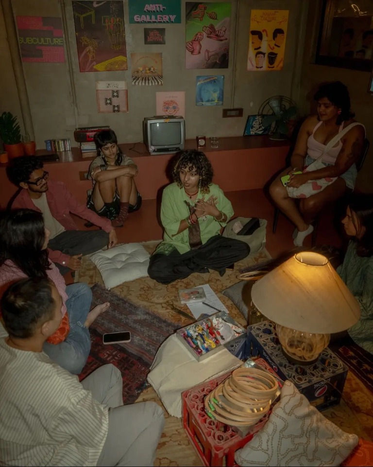 A queer community gathered in a warmly lit room, sitting together in conversation.
